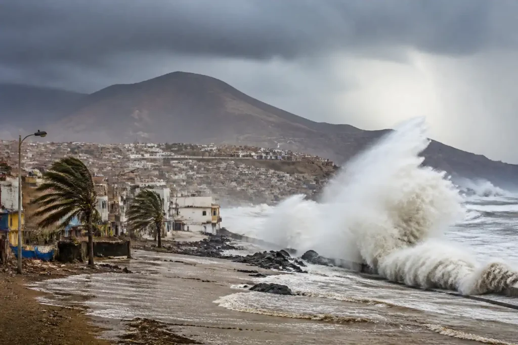 Stormy waves crash onto a seaside promenade with mountains and palm trees in the background